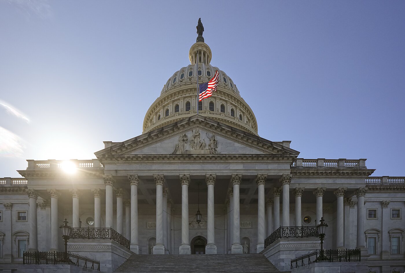 U.S. Capitol with sun behind the dome and an American flag flying against a clear blue sky.